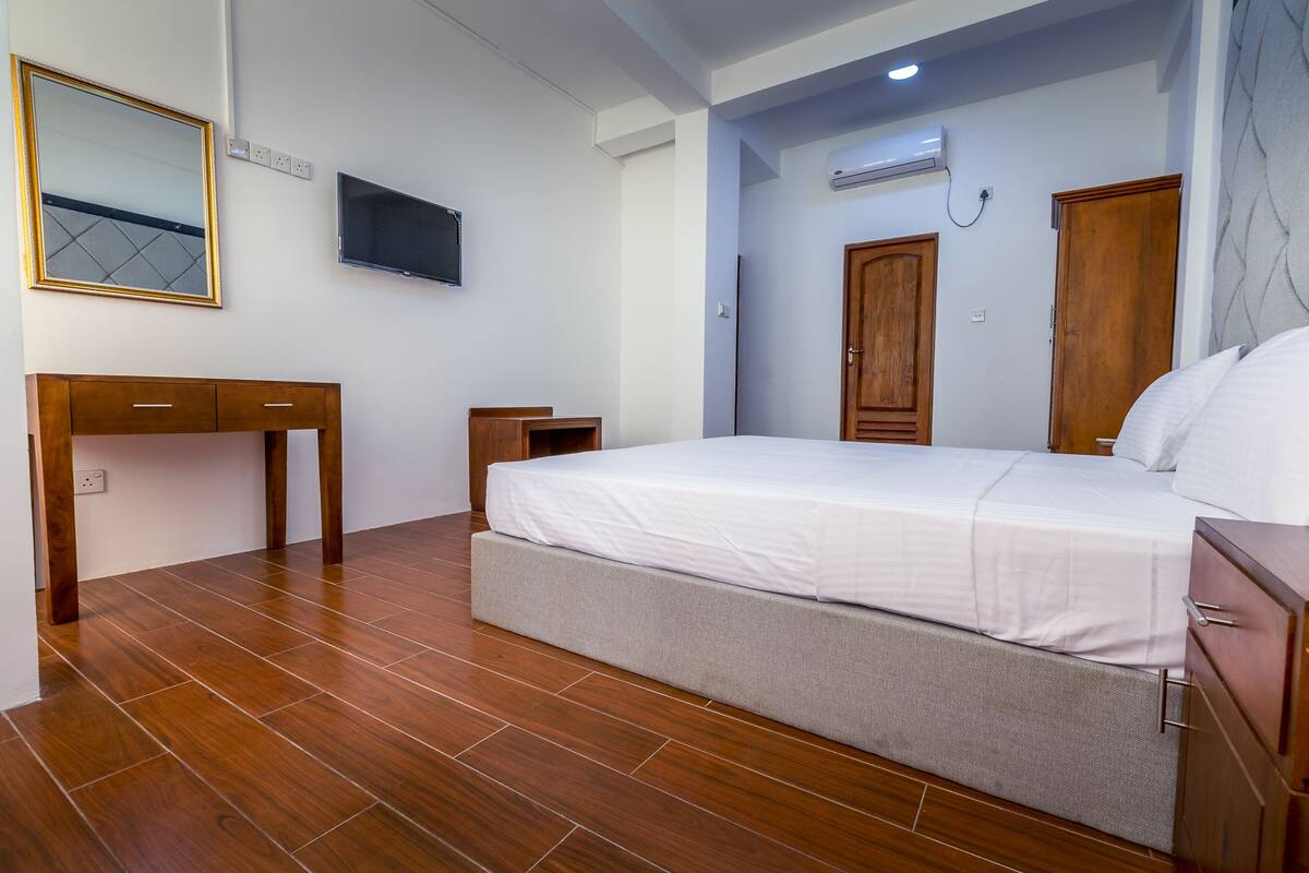 A bedroom with a bed, a desk, a television, and an air conditioning unit.