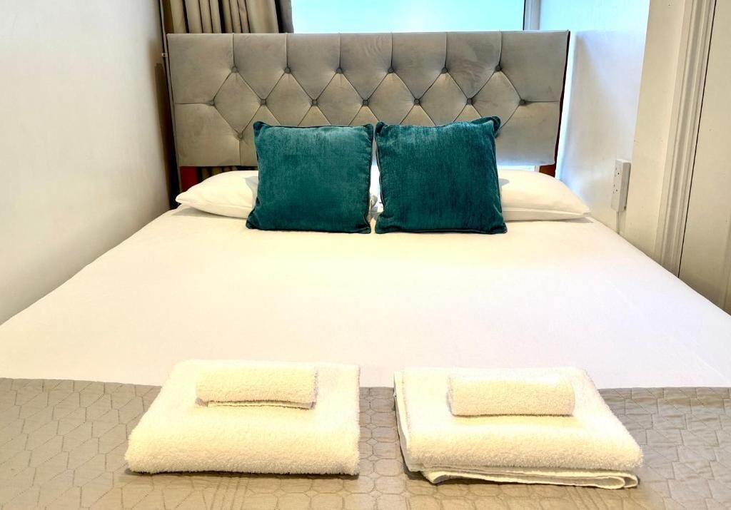 A bed with a tufted headboard, two teal pillows, and two folded white towels on a patterned carpet.
