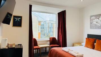 Quarto de hotel com uma cama, duas cadeiras, uma escrivaninha, janela com cortinas e obras de arte nas paredes.