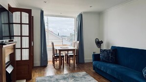 A modern living room with a dining area, a blue sofa, and a view of the cityscape through large windows. - Reef Lodge (Newquay)