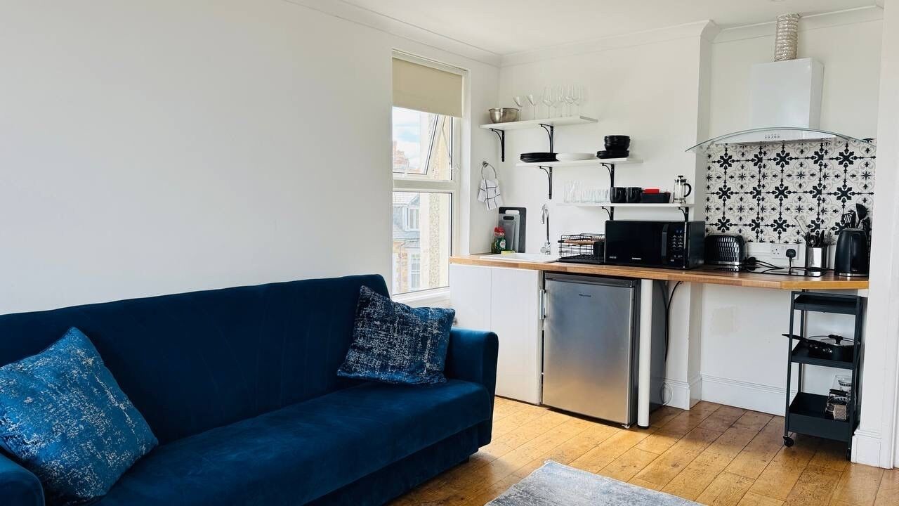 Un espacio habitable compacto con kitchenette, un sofá azul con cojines estampados y una ventana que proporciona luz natural.