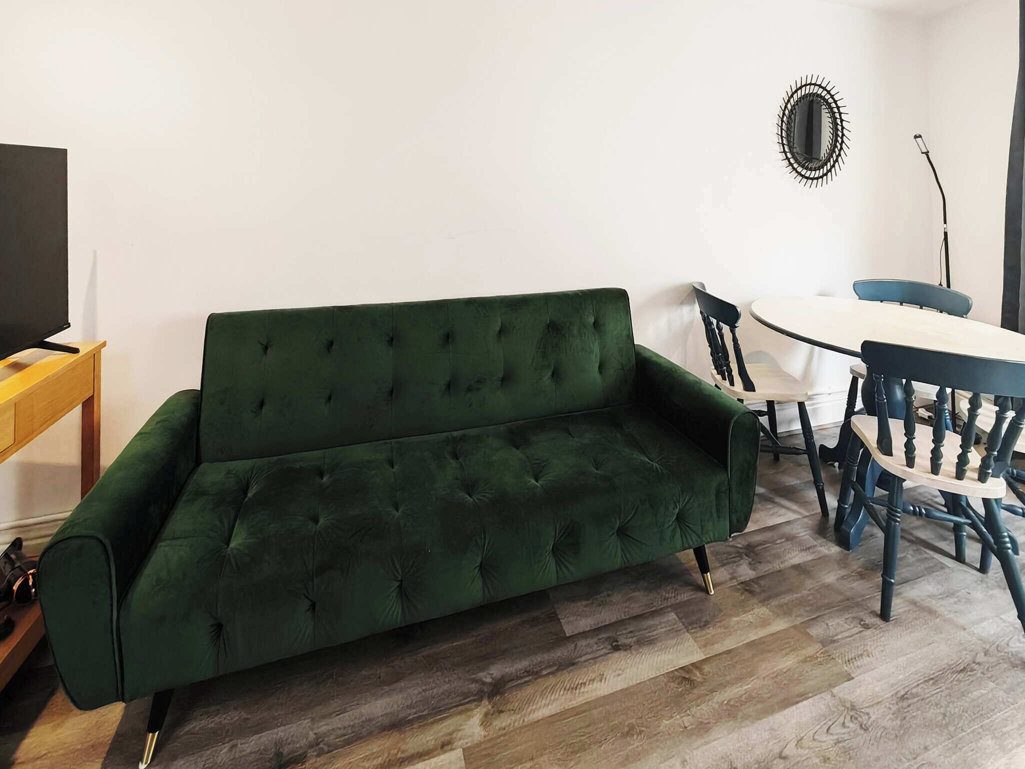 A green sofa, a wooden TV stand, a dining table with chairs, and a mirror on the wall.