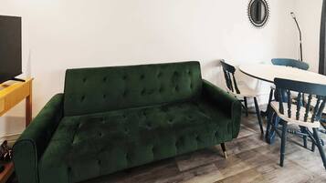 A green sofa, a wooden TV stand, a dining table with chairs, and a mirror on the wall.