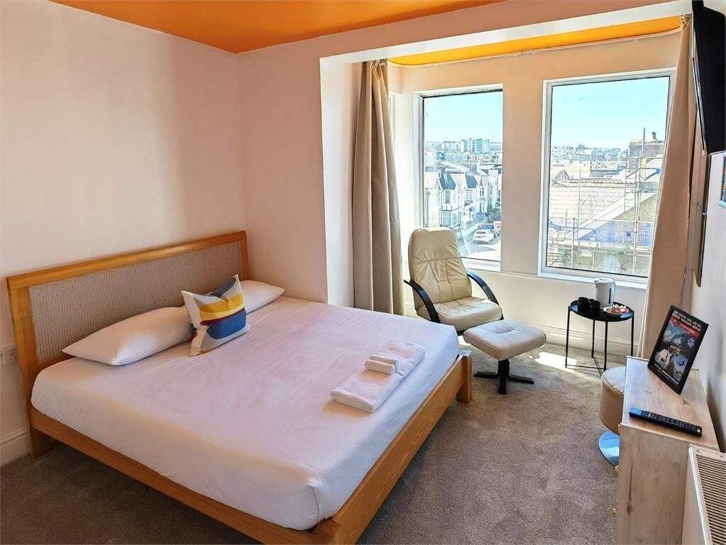 A hotel room with a bed, a chair, a small table, a window with a city view, and a picture frame on a desk.