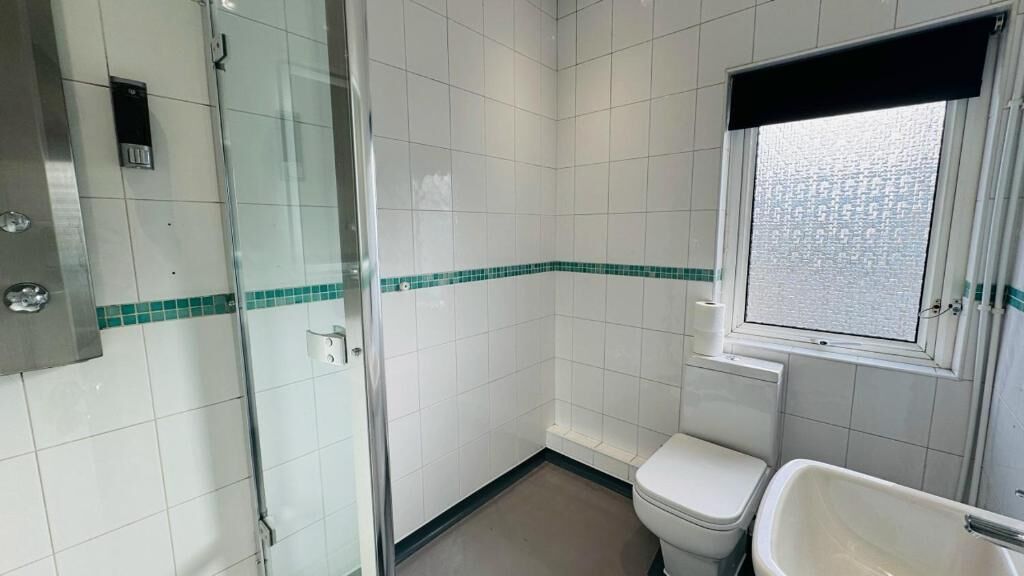 A bathroom with white tiles, a glass shower enclosure, a toilet, and a window.