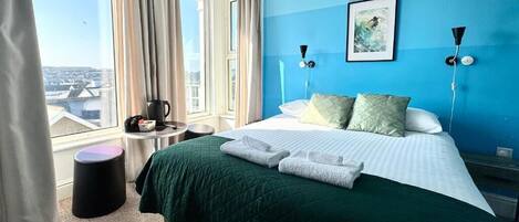 Hotelkamer met een bed, uitzicht op de stad, een raam met gordijnen en een klein tafeltje met waterkoker.