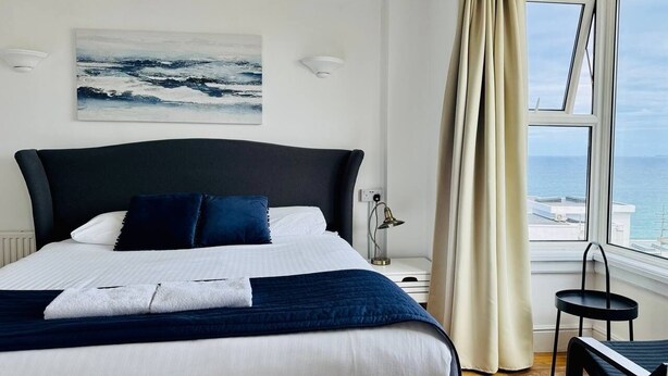 A neatly made bed with a dark headboard, a bedside lamp, and a view of the sea through a window with curtains. - Reef Lodge (Newquay)