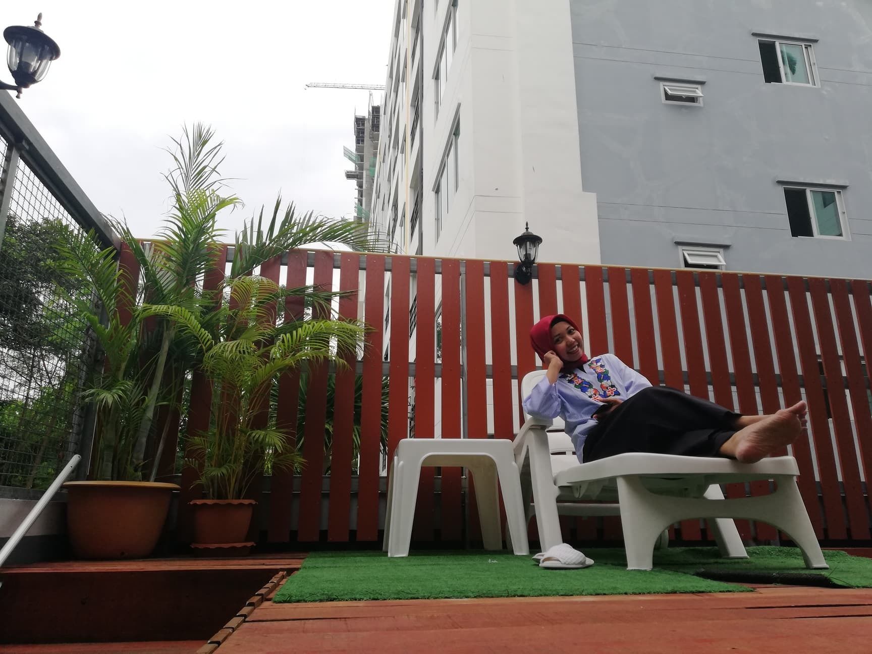 A person sitting on a white plastic chair in an outdoor area with a red fence, potted plants, and a building in the background.