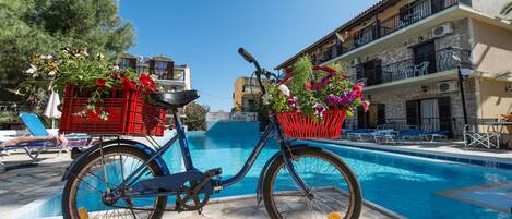A blue bicycle with baskets containing flowers, parked by a poolside with lounge chairs and a building in the background.