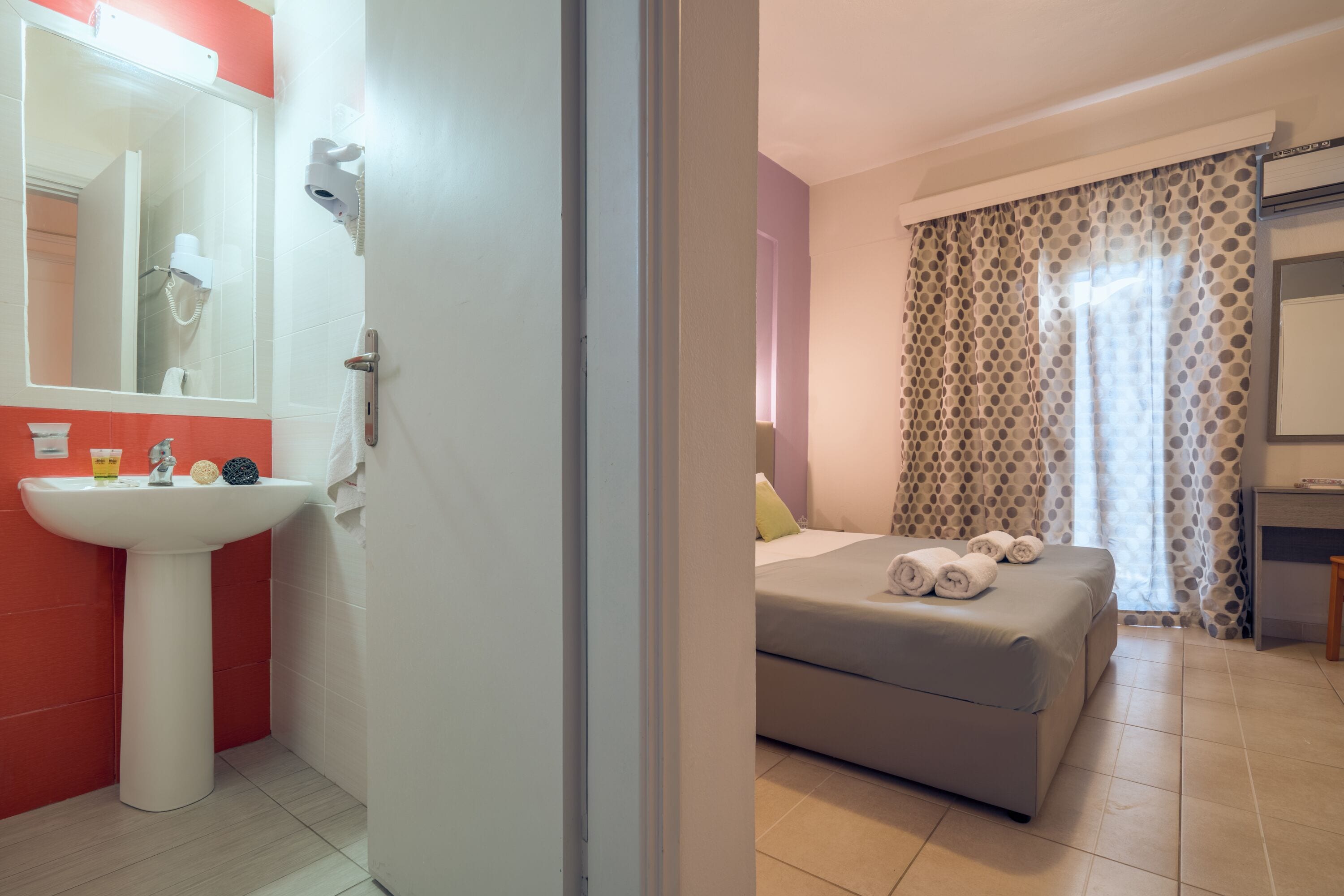 A hotel room with a bathroom featuring a sink, mirror, and hairdryer. The room has a bed with a pillow and rolled towels.