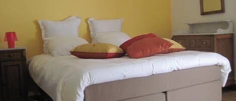 A bed with white and red pillows, a wooden nightstand with a red object, and a mirror on the wall.