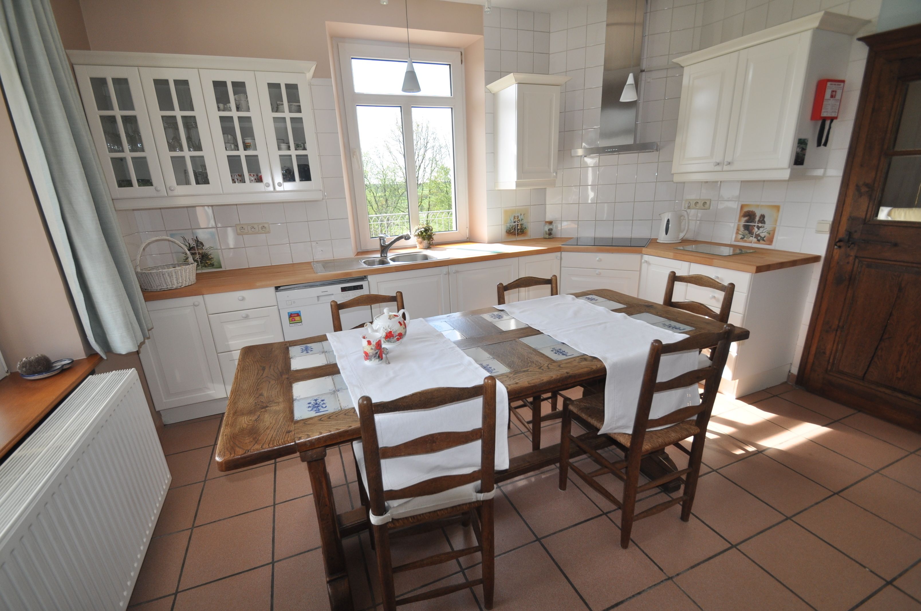 A kitchen with white cabinets, a wooden table, and chairs. There is a window with a view of trees outside.