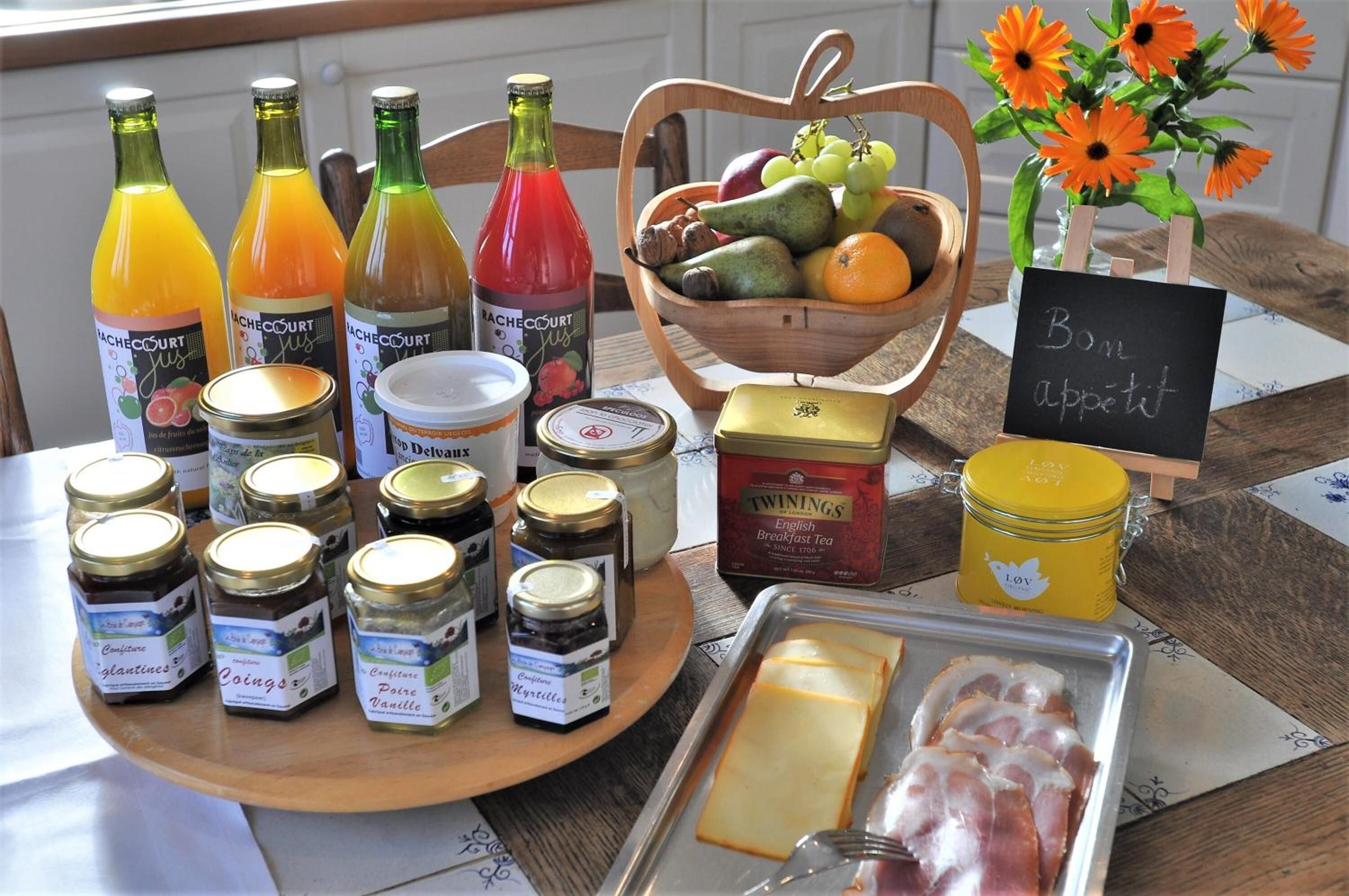 Ein Frühstückstisch mit verschiedenen Speisen, darunter Gläser mit Marmelade, ein Obstkorb, eine Käseplatte und eine Saftflasche.