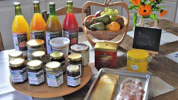 Ein Frühstückstisch mit verschiedenen Speisen, darunter Gläser mit Marmelade, ein Obstkorb, eine Käseplatte und eine Saftflasche.