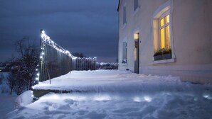 A snow-covered pathway leading to a building with a lit window and a fence with lights. - B&B Le Verger du Pierroy (Léglise)