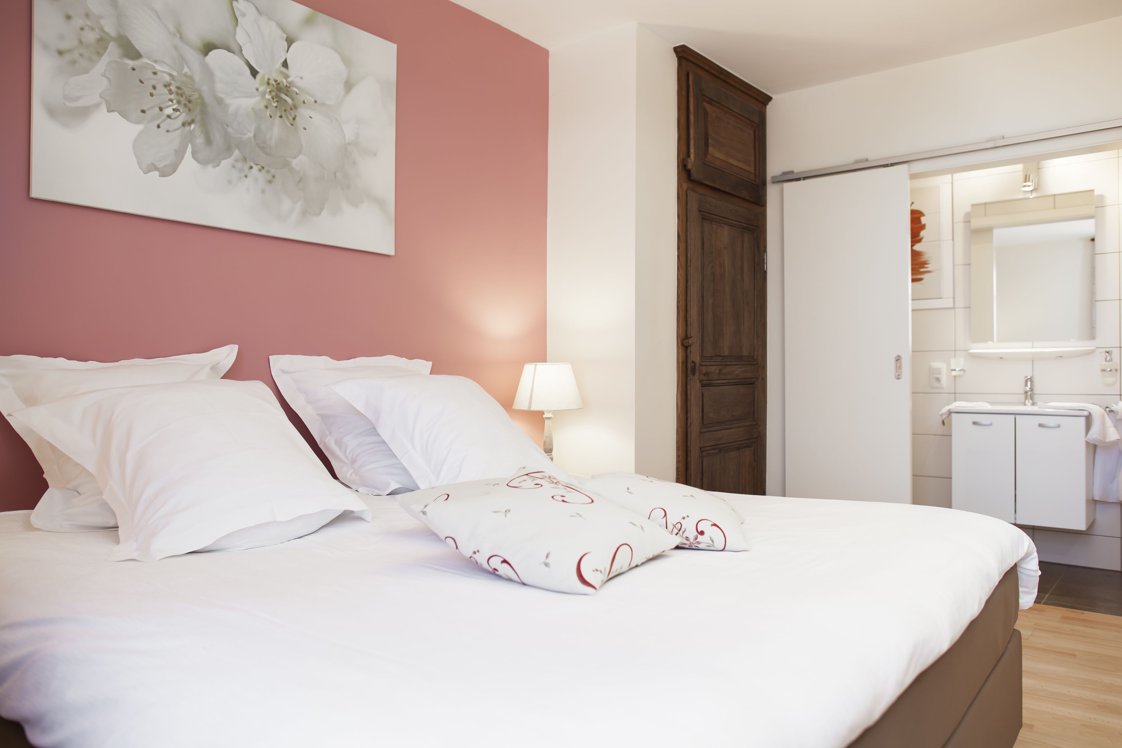 A neatly made bed with white linens and decorative pillows, a bedside lamp, a wooden wardrobe, and a bathroom with a mirror and sink.