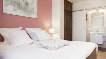 A neatly made bed with white linens and decorative pillows, a bedside lamp, a wooden wardrobe, and a bathroom with a mirror and sink.
