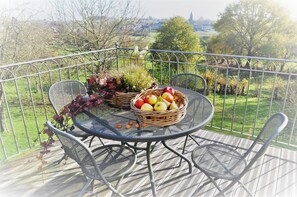 A balcony with a round table and two chairs, a wicker basket with fruits, and a view of a green landscape. - B&B Le Verger du Pierroy (Léglise)