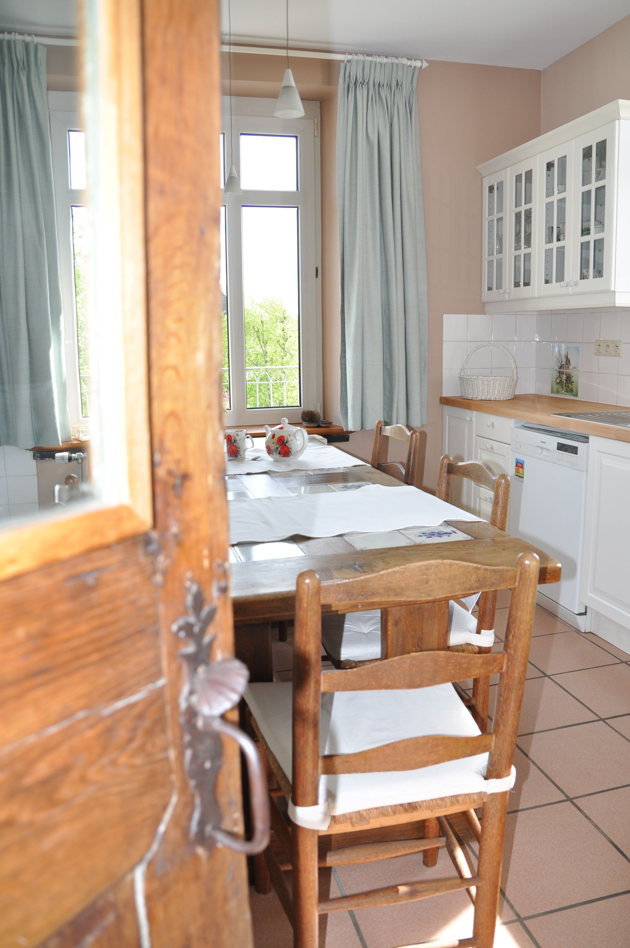 Zona pranzo con tavolo e sedie in legno, cucina con mobili bianchi e finestra con tende.