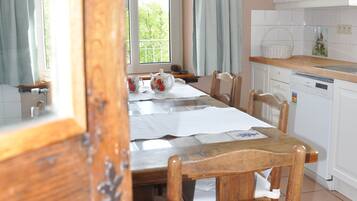 Zona pranzo con tavolo e sedie in legno, cucina con mobili bianchi e finestra con tende.