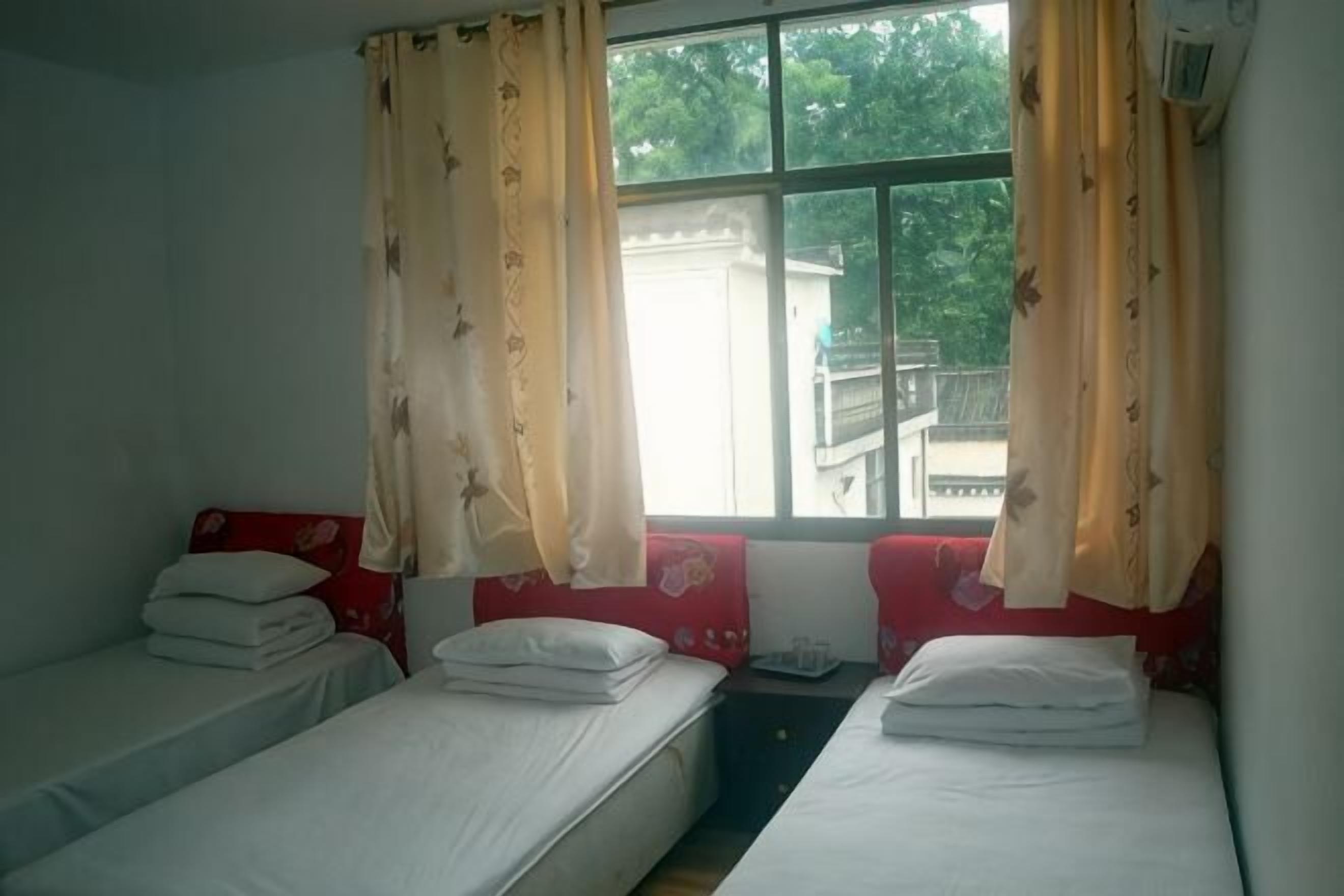 A room with two single beds, a window with curtains, and a view of greenery outside.