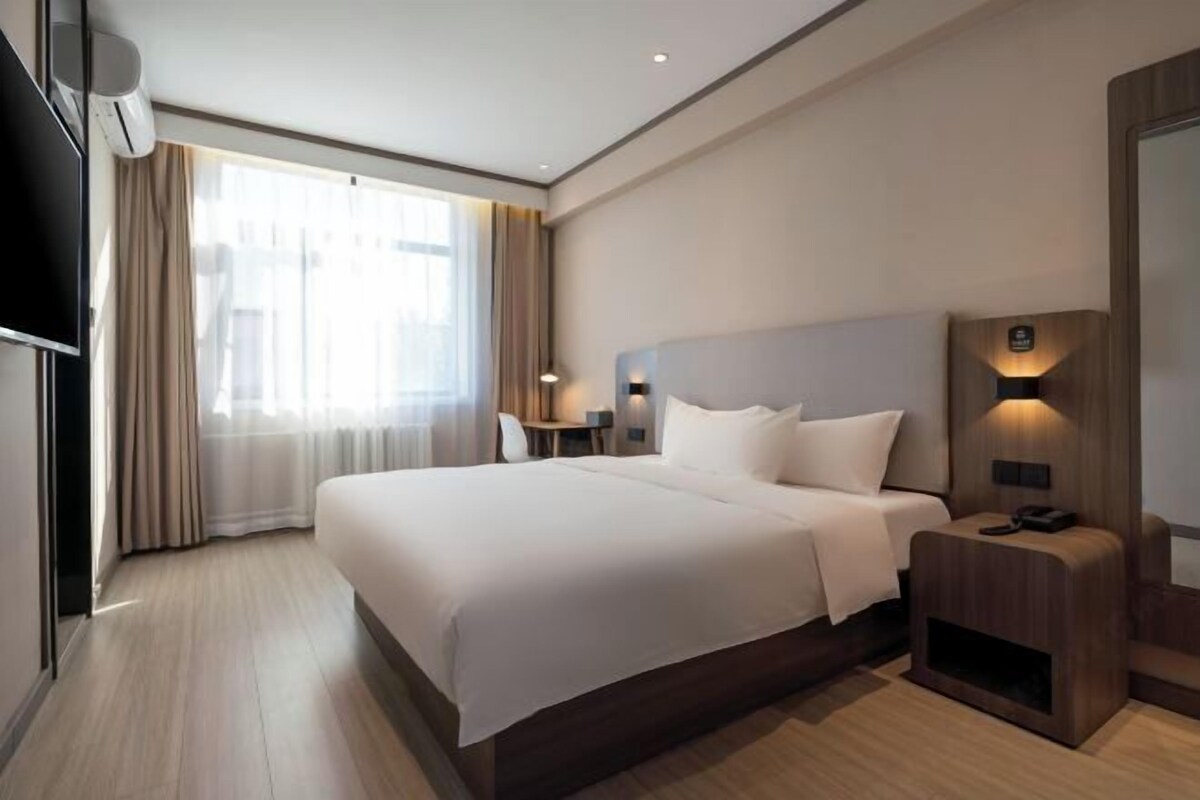 A modern hotel room with a large bed, a bedside table with a lamp, a flat-screen TV, and a large window with curtains.