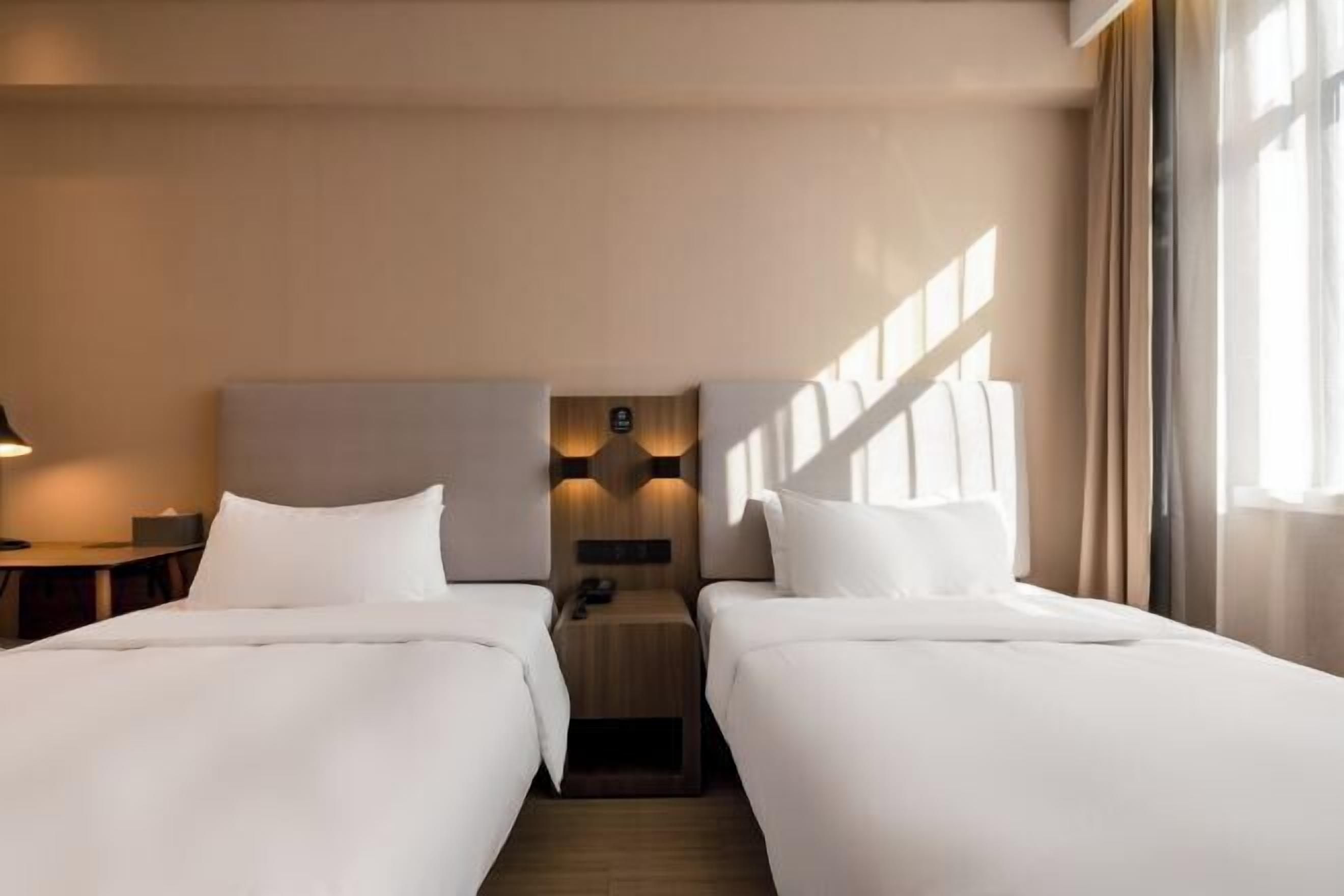 Habitación de hotel con dos camas, una mesita de noche, una lámpara de pared y un ventanal con cortinas.