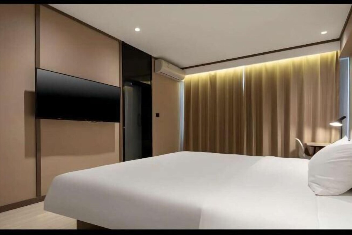 A hotel room with a bed, a flat-screen TV mounted on the wall, a bedside table with a lamp, and a curtain-covered window.