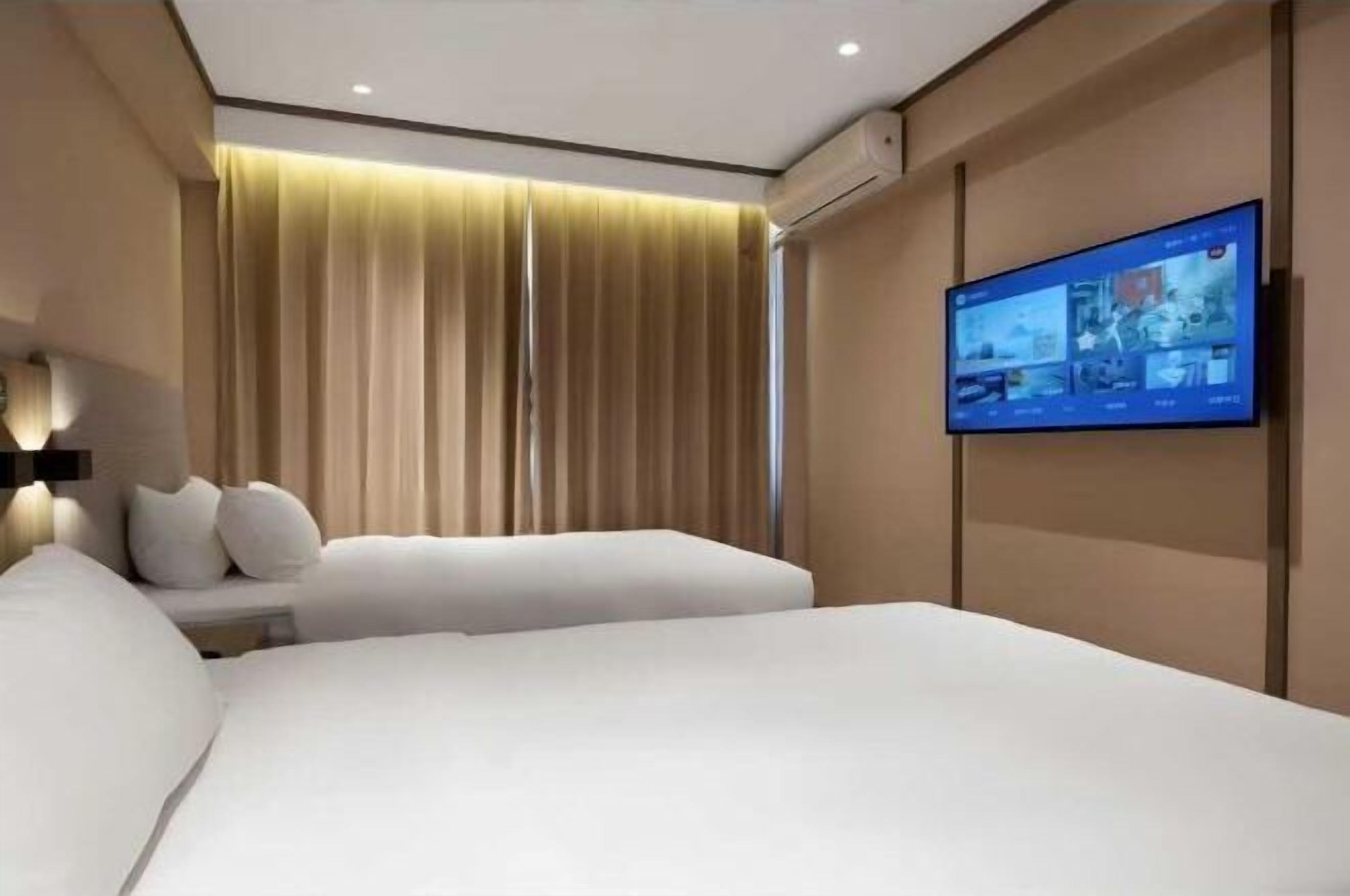 A modern hotel room with a large bed, two bedside tables, a flat-screen TV mounted on the wall, and a large window with curtains.