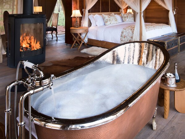 A bathroom with a freestanding bathtub, a wood-burning stove, and a bed with a canopy. - One Nature Nyaruswiga Serengeti (Serengeti)