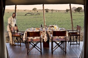 An outdoor dining area with a table set for four, surrounded by wooden chairs, and a person standing to the side. - One Nature Nyaruswiga Serengeti (Serengeti)