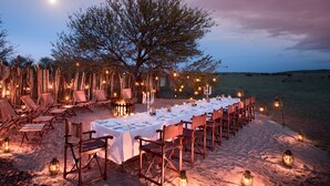An outdoor dining setup with a long table, chairs, and lanterns, surrounded by a wooden fence and a tree, set in a sandy area. - One Nature Nyaruswiga Serengeti (Serengeti)