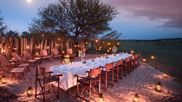 An outdoor dining setup with a long table, chairs, and lanterns, surrounded by a wooden fence and a tree, set in a sandy area.