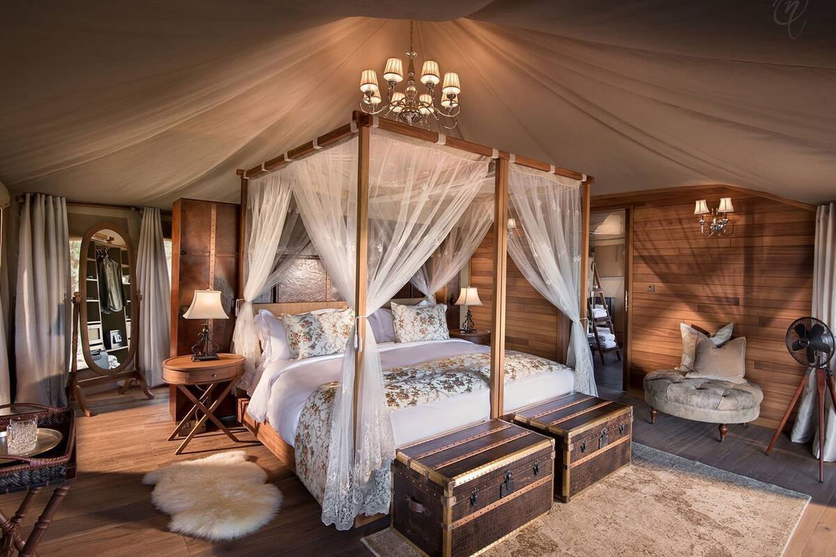 A four-poster canopy bed with a wooden finish, bedside tables with lamps, a fluffy white rug, and a vintage-style armchair.