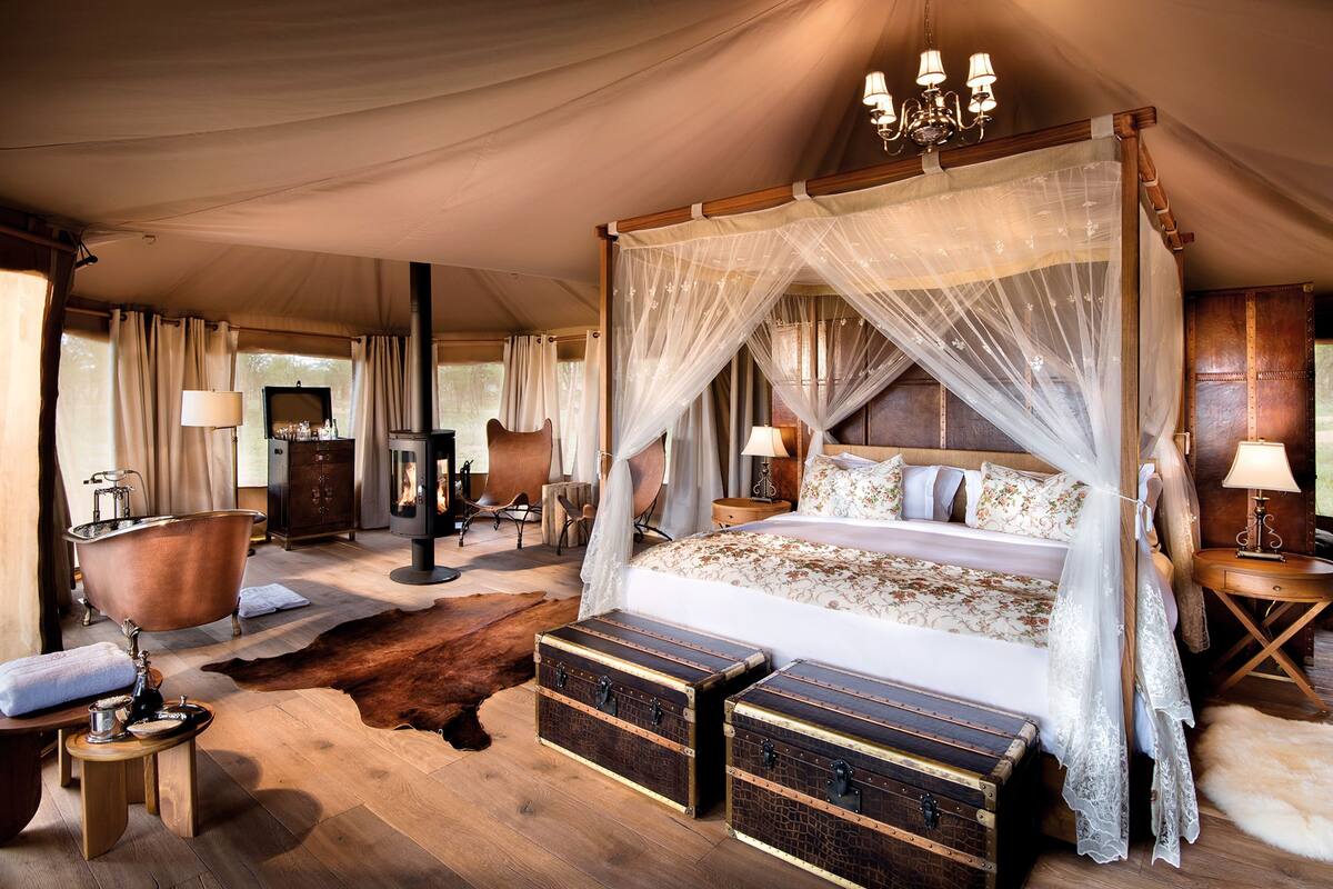 A spacious tented room with a four-poster bed, a wood-burning stove, leather furniture, and a large fur rug.