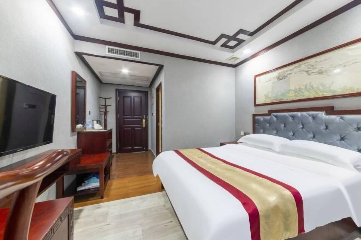 A hotel room with a bed, a TV, a desk, and a painting on the wall.