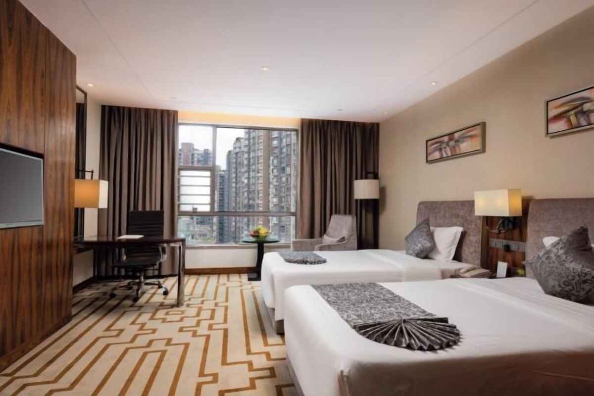 A hotel room with two beds, a desk, a chair, a TV, and a large window with a city view.