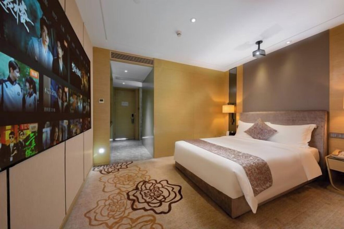 A hotel room with a large bed, a wall-mounted TV displaying movie posters, and a carpet with a floral pattern.