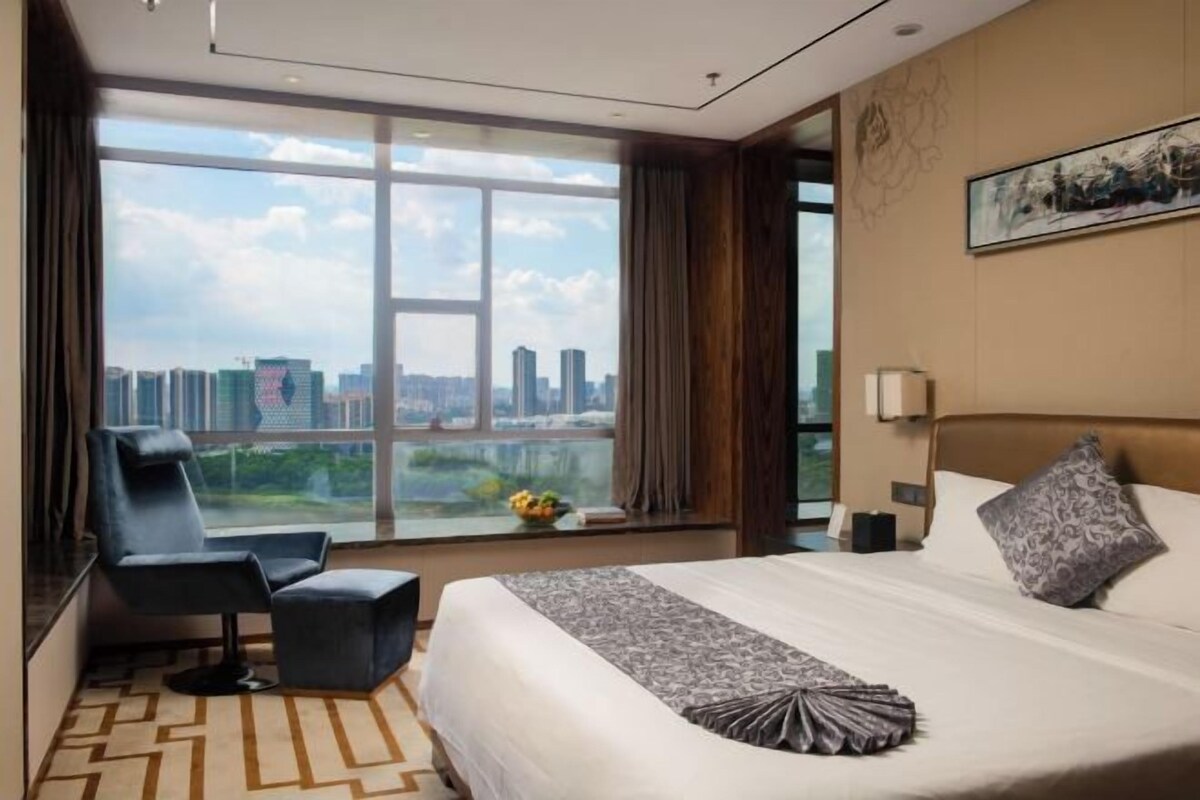 A hotel room with a large window offering a city view, a bed with a patterned throw, a chair, and a small table with a vase of flowers.