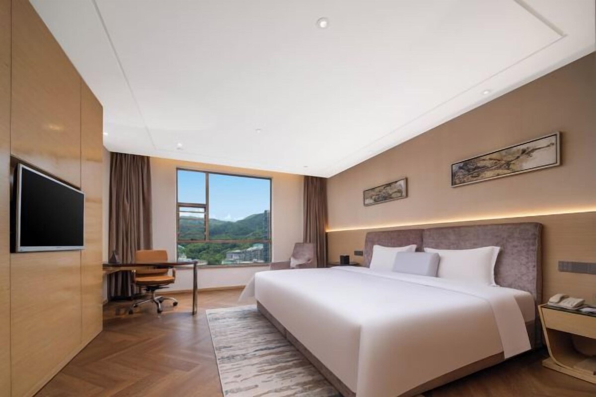 A modern hotel room with a large bed, a desk, a chair, a TV, and a window with a view of the mountains.