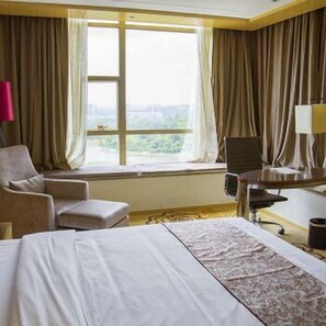 A hotel room with a bed, two armchairs, a desk with a chair, a window with curtains, and a view of greenery outside. - City Hotel (Shenzhen)