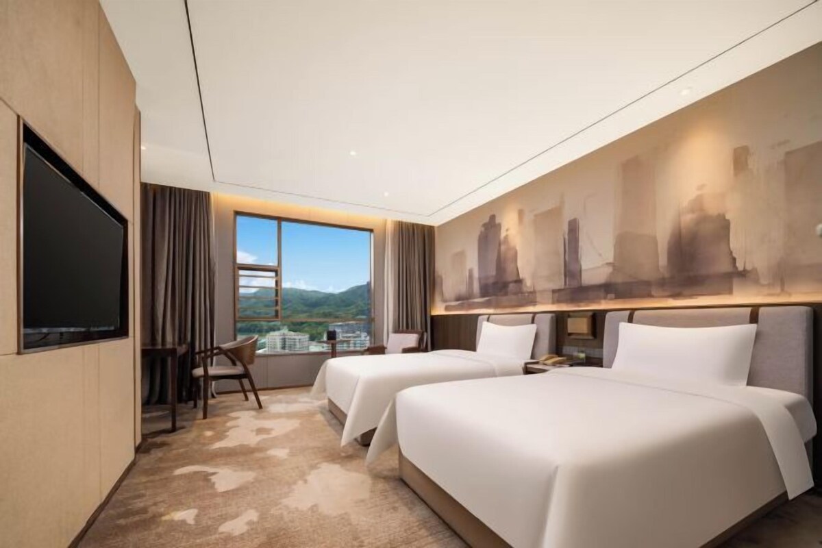 A hotel room with two beds, a large TV, a view of the city, and a painting on the wall.