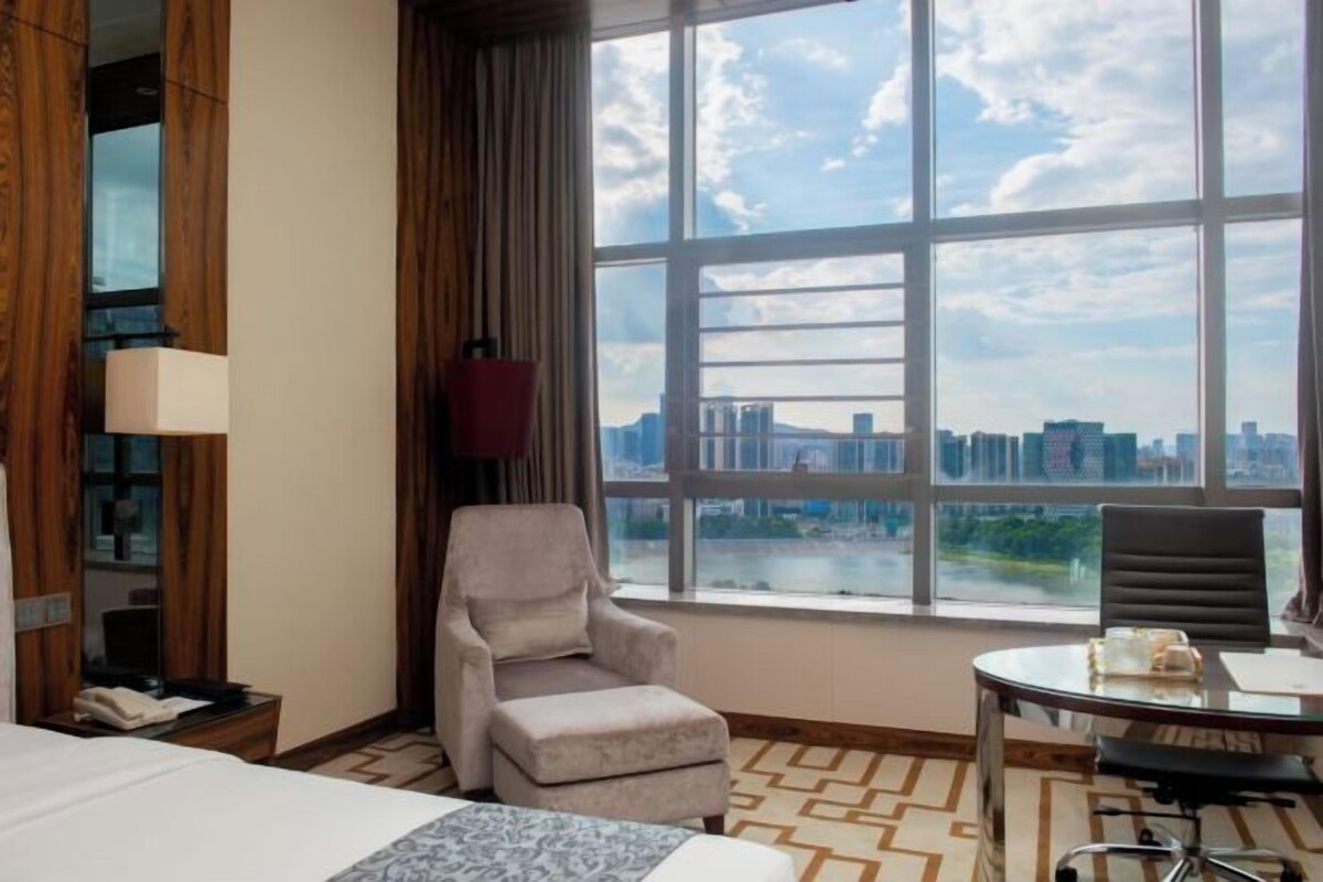 A hotel room with a large window offering a city view, a bed, a desk with a chair, and a small table with a lamp.