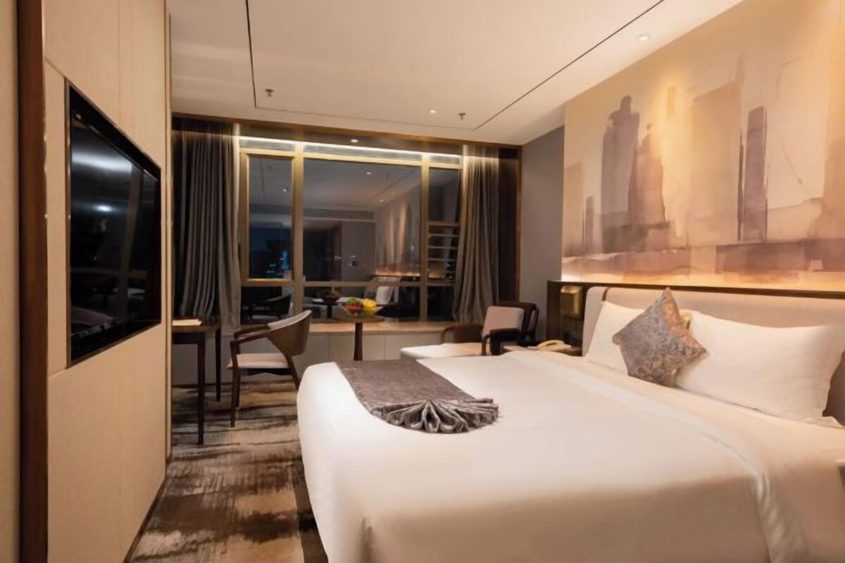 A hotel room with a large bed, a desk with a chair, a view of the city through large windows, and a painting on the wall.