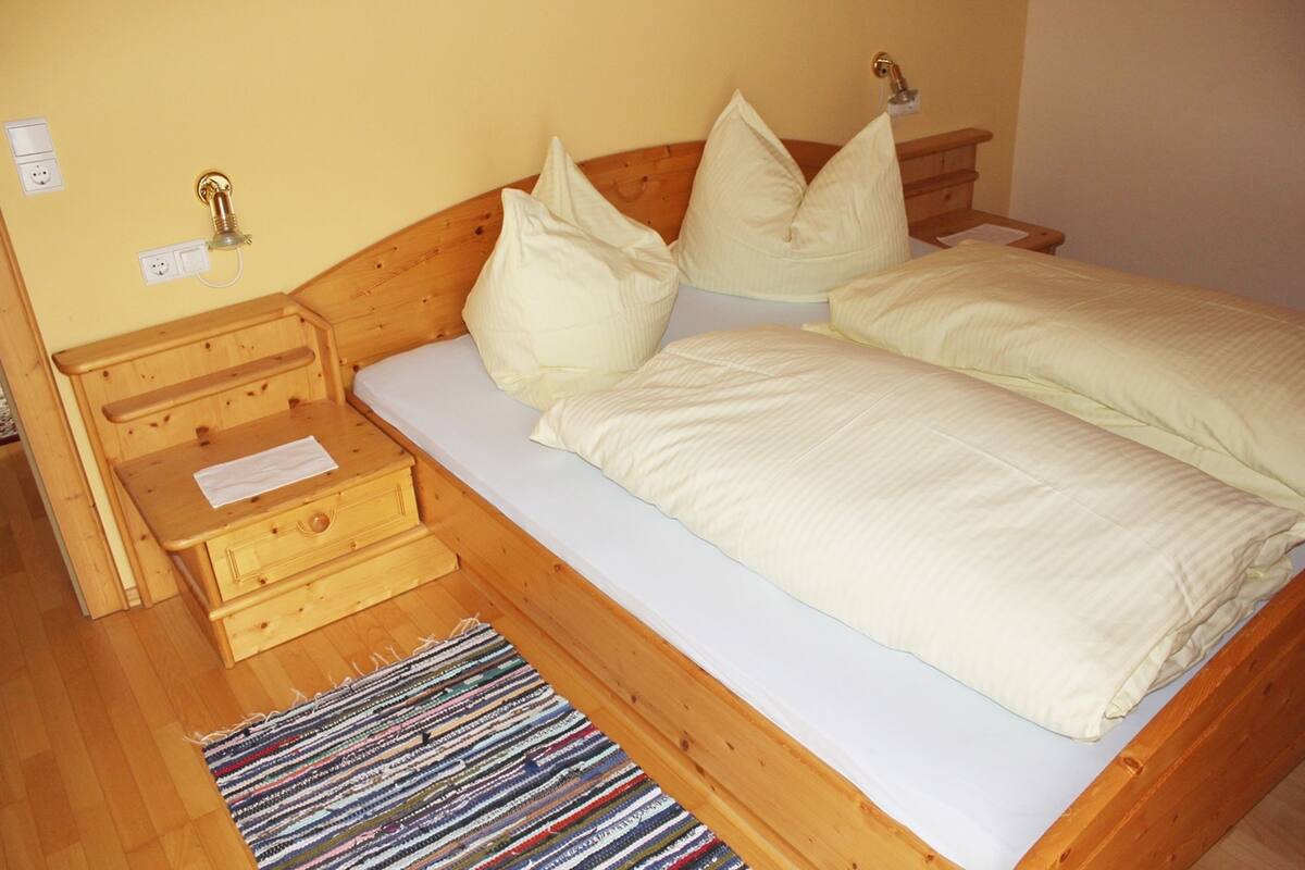 Ein hölzernes Bett mit weißer Bettwäsche und Kissen, ein Nachttisch mit Lampe sowie eine kleine Holzschatulle.