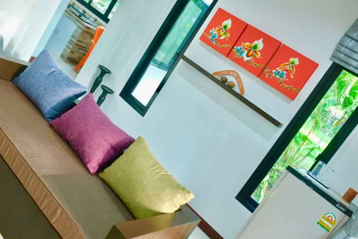 Uma sala de estar moderna com um sofá, almofadas coloridas, um objeto decorativo na parede e vista para o verde pela janela.
