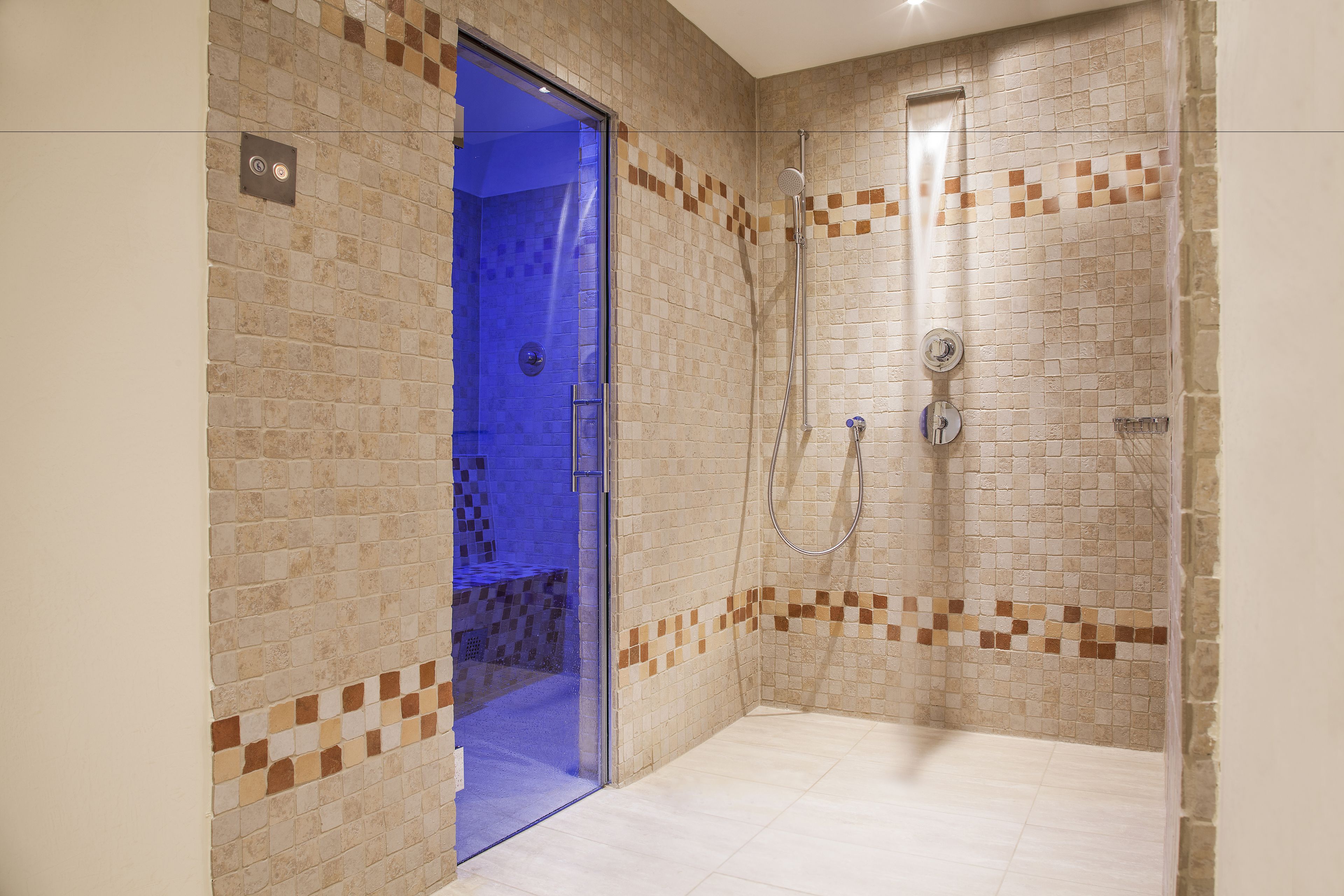 Un espacio de ducha con paredes y suelo de azulejos, una puerta de cristal que da al baño y un cabezal de ducha en la pared.