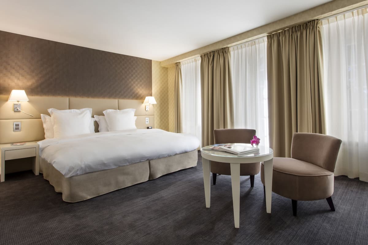 Habitación de hotel con una cama grande, dos mesitas de noche con lámparas, una mesa redonda con una silla y vistas al exterior a través de cortinas translúcidas.