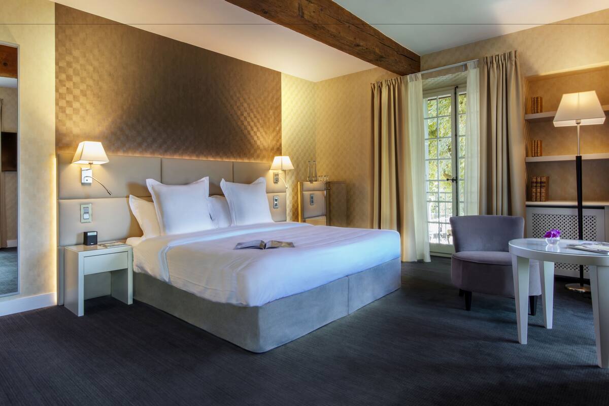 Una habitación de hotel moderna con una cama grande, dos mesitas de noche con lámparas, una pequeña mesa con una silla y vistas al exterior a través de una ventana.