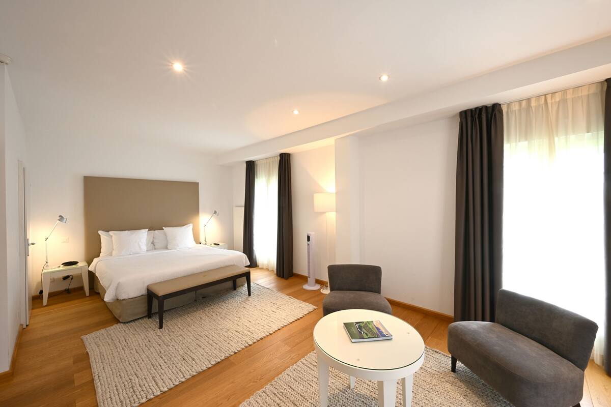 Una habitación de hotel con cama, mesita de noche, lámpara, silla y una pequeña mesa con un libro.